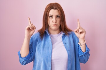 Young hispanic woman with red hair standing over pink background pointing up looking sad and upset,...