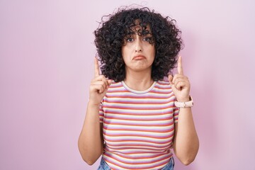 Young middle east woman standing over pink background pointing up looking sad and upset, indicating...