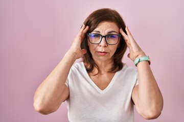 Middle age hispanic woman standing over pink background with hand on head, headache because stress. suffering migraine.