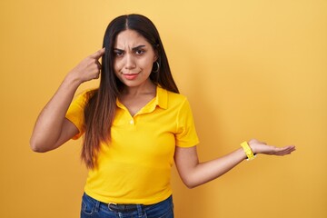 Young arab woman standing over yellow background confused and annoyed with open palm showing copy...