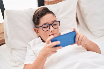 Obraz na płótnie Canvas Adorable hispanic boy watching video on smartphone lying on bed at bedroom