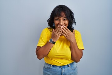 Hispanic woman standing over blue background laughing and embarrassed giggle covering mouth with...