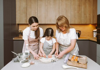 Obraz na płótnie Canvas Smiling three generations of women have fun making dough in the kitchen. A happy little girl with her mother and older grandmother are preparing pastries or cookies.