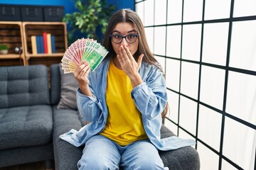 Young hispanic woman holding norwegian banknotes covering mouth with hand, shocked and afraid for...
