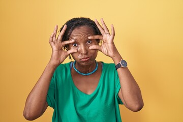 African woman with dreadlocks standing over yellow background trying to open eyes with fingers, sleepy and tired for morning fatigue