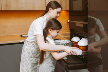 A little girl and her mother put a form with Easter cakes in the oven in the kitchen. Mom and child...