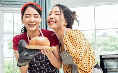Asian beautiful adorable two women or friends wearing aprons, closing eyes and smiling with happiness, smelling good taste while holding dish of baked bread in kitchen at comfortable home on weekend.