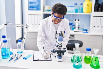 African american woman wearing scientist uniform using microscope write on clipboard at laboratory