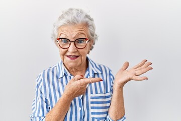 Senior woman with grey hair standing over white background amazed and smiling to the camera while presenting with hand and pointing with finger.