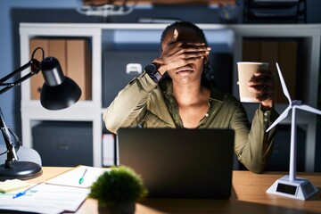 African woman working using computer laptop at night covering eyes with hand, looking serious and...