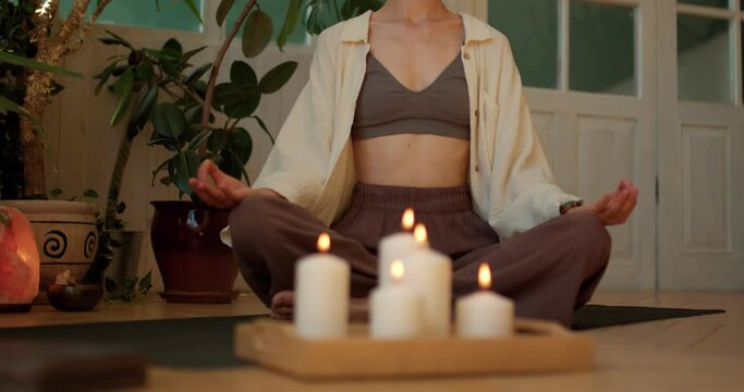 slim relaxed woman doing yoga sits in lotus position in front of candles,female meditates at home sitting on floor zoom out motion,mindfulness spirituality concept,blurred foreground slow motion