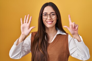 Young brunette woman standing over yellow background wearing glasses showing and pointing up with fingers number seven while smiling confident and happy.