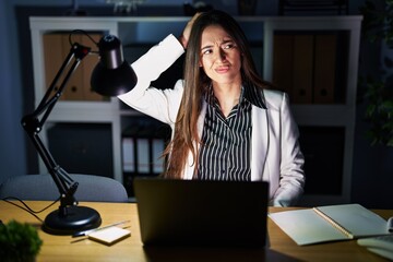 Young brunette woman working at the office at night with laptop confuse and wondering about question. uncertain with doubt, thinking with hand on head. pensive concept.
