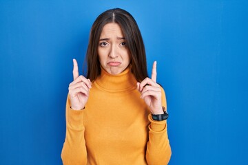 Young brunette woman standing over blue background pointing up looking sad and upset, indicating...