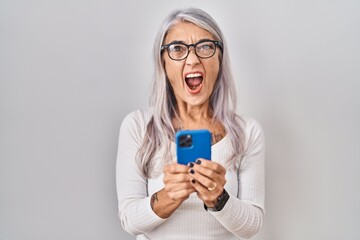 Middle age woman with grey hair using smartphone typing message angry and mad screaming frustrated and furious, shouting with anger. rage and aggressive concept.