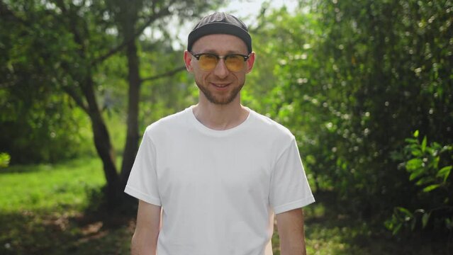 Portrait of authentic man looking at camera and smiling outdoor. Handsome bearded guy in white T-shirt and yellow glasses posing on camera outside