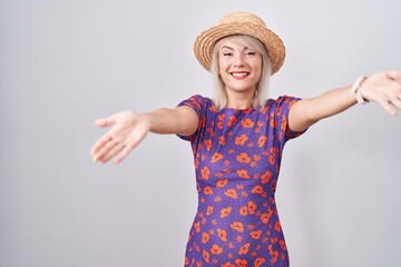 Young caucasian woman wearing flowers dress and summer hat looking at the camera smiling with open arms for hug. cheerful expression embracing happiness.