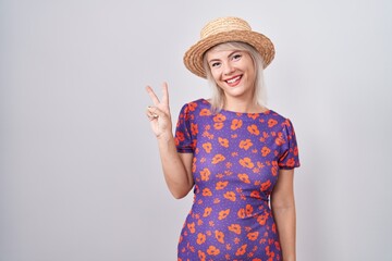 Young caucasian woman wearing flowers dress and summer hat showing and pointing up with fingers number two while smiling confident and happy.
