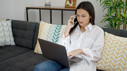 Young beautiful hispanic woman talking on smartphone using laptop at home