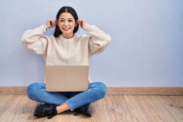 Young woman using laptop sitting on the floor at home smiling pulling ears with fingers, funny...