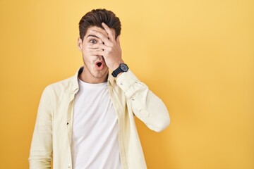 Young hispanic man standing over yellow background peeking in shock covering face and eyes with hand, looking through fingers with embarrassed expression.