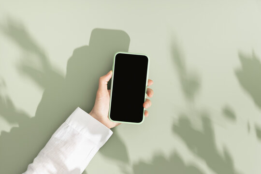 Woman hold phone with black blank screen, minimal flat lay on olive green background. Smartphone mock up. Digital online summer lifestyle composition with leaves shadows and sunlight