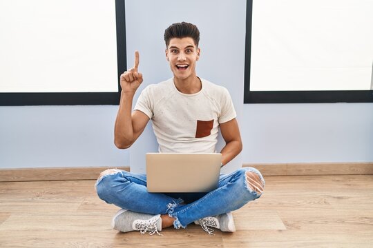 Young hispanic man using laptop at home pointing finger up with successful idea. exited and happy. number one.