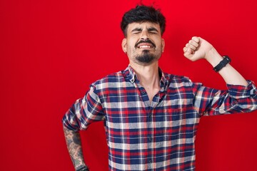 Fototapeta premium Young hispanic man with beard standing over red background stretching back, tired and relaxed, sleepy and yawning for early morning