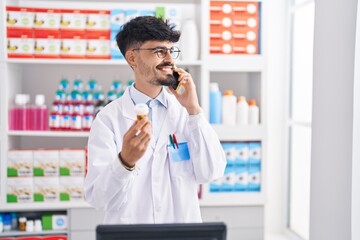Young hispanic man pharmacist talking on smartphone holding pills at pharmacy