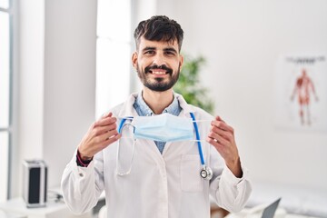 Young hispanic doctor man with beard holding safety mask smiling with a happy and cool smile on face. showing teeth.