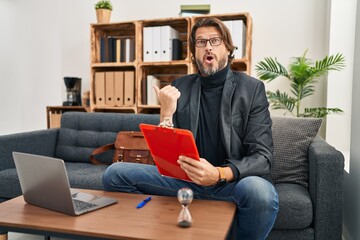 Fototapeta premium Handsome middle age man working at consultation office surprised pointing with hand finger to the side, open mouth amazed expression.