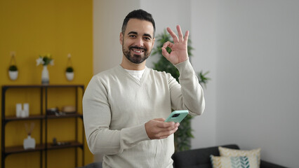 Young hispanic man using smartphone doing ok gesture at home