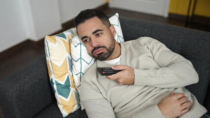 Young hispanic man watching tv lying on sofa with boring expression at home