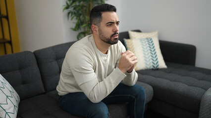 Young hispanic man sitting on sofa with worry expression at home