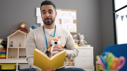 Young hispanic man preschool teacher reading story book sitting on table at kindergarten