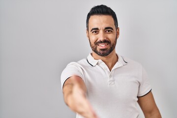 Young hispanic man with beard wearing casual clothes over white background smiling friendly offering handshake as greeting and welcoming. successful business.