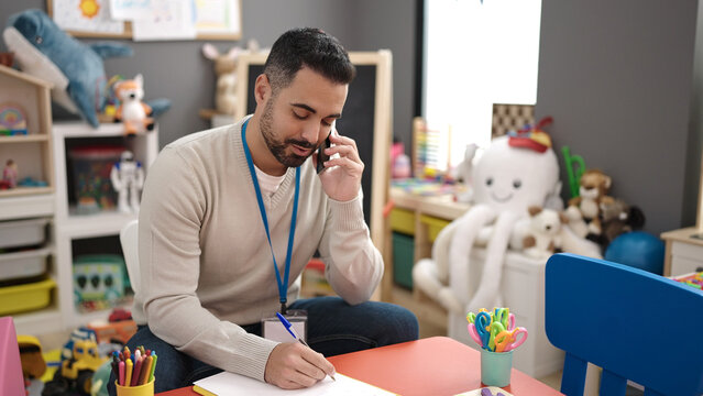 Young hispanic man preschool teacher writing on document talking on smartphone at kindergarten
