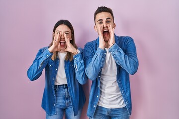 Young hispanic couple standing over pink background shouting angry out loud with hands over mouth