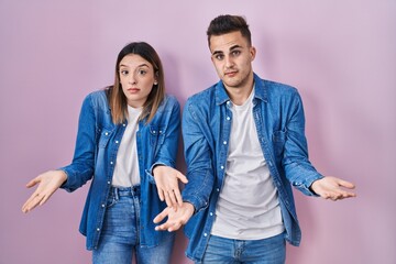 Young hispanic couple standing over pink background clueless and confused with open arms, no idea...