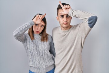 Young hispanic couple standing over white background making fun of people with fingers on forehead...
