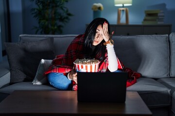 Hispanic woman eating popcorn watching a movie on the sofa surprised with hand on head for mistake,...