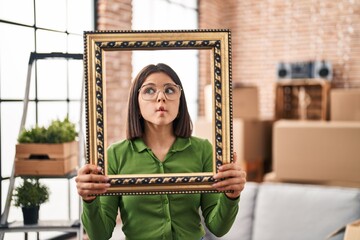 Young hispanic woman at new home holding empty frame making fish face with mouth and squinting eyes, crazy and comical.