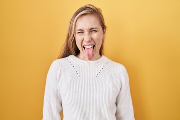 Young caucasian woman wearing white sweater over yellow background sticking tongue out happy with...