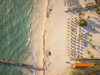 Wooden pier in the sea. View from above. Lots of beach umbrellas on the sandy beach, palm trees, boats. Seaside resort, recreation, boat trips, fishing, relaxation.