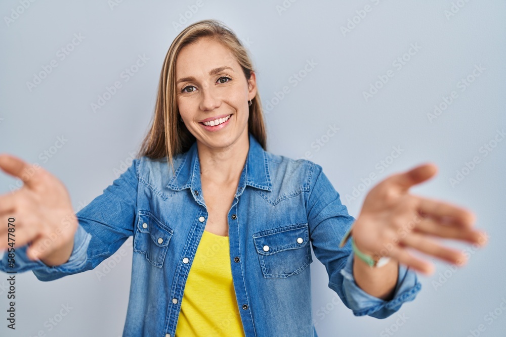 Poster Young blonde woman standing over blue background smiling cheerful offering hands giving assistance and acceptance.