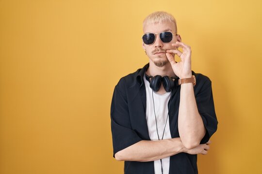 Young caucasian man wearing sunglasses standing over yellow background mouth and lips shut as zip with fingers. secret and silent, taboo talking