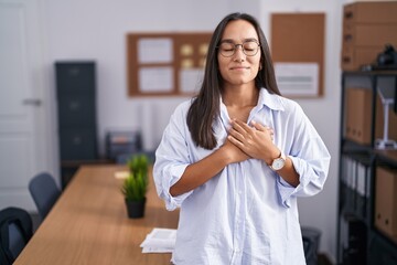 Plakat Young hispanic woman at the office smiling with hands on chest with closed eyes and grateful gesture on face. health concept.