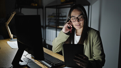 Young beautiful hispanic woman business worker using tablet talking on smartphone at office