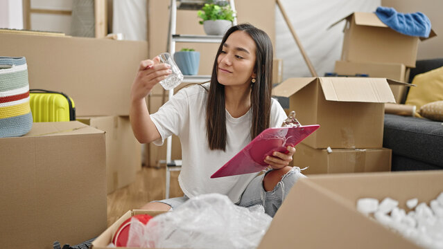 Young beautiful hispanic woman unpacking cardboard box checking on checklist at new home