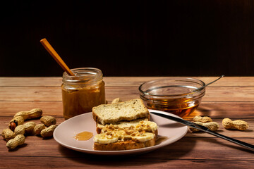 Peanut butter or peanut paste in an open jar and spread on a toast sandwich with banana and honey and peanuts in shells scattered on the table